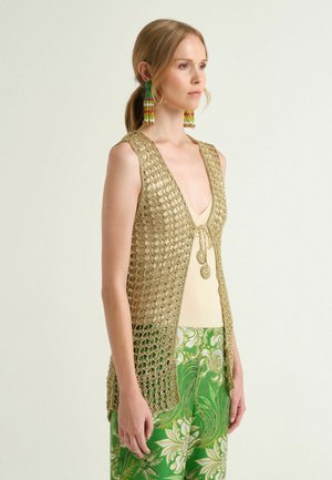 Femme portant un gilet sans manches beige en maille ouverte sur un haut crème et un pantalon vert à motifs floraux, avec de longues boucles d'oreilles multicolores en perles.