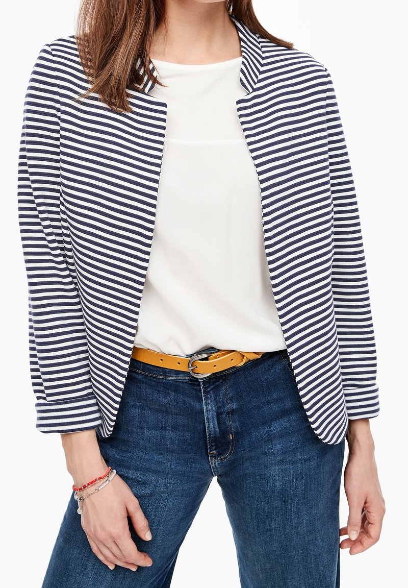 Blazer rayé en marine et blanc, associé à un haut blanc et un jean bleu. Accentué par une ceinture jaune. Tissu lisse, design décontracté.