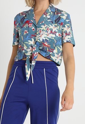 Femme portant une chemise courte à manches courtes, bleue à fleurs, nouée devant, associée à un pantalon taille haute bleu foncé avec des rayures verticales blanches.