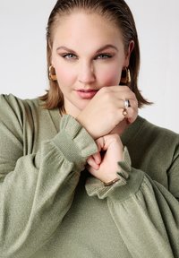 Groen, gestructureerd gebreide trui met opgeblazen mouwen en gerimpelde manchetten. Accessoires omvatten gouden oorringen en meerdere ringen.