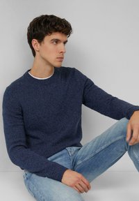 Granatowy sweter o fakturze, z prążkowanymi mankietami i dekoltem, w połączeniu z jasnobłękitnymi dżinsami. Model siedzi na neutralnym tle.