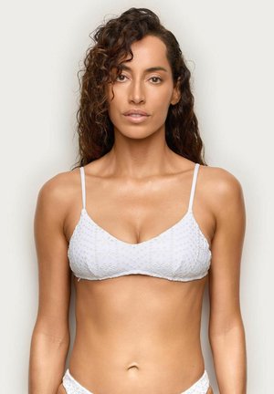 Kobieta o kręconych brązowych włosach, ubrana w biały, teksturowany top bikini i pasujące doły, stojąca na jednolitym tle.