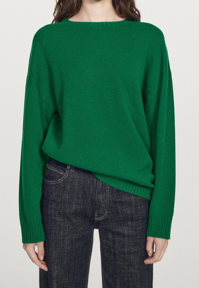 Personne portant un pull vert près du corps à manches longues et un jean en denim bleu foncé, debout devant un fond clair uni.