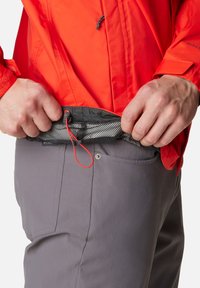 Rote Jacke mit strukturiertem Finish, kombiniert mit grauen Hosen, die über eine Netztasche und einen wieder sichtbaren roten Anpassungskordel am Bund verfügen.