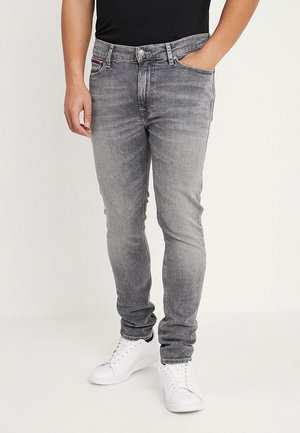 Homme portant un jean skinny gris délavé, les mains dans les poches et des baskets blanches sur un fond uni.