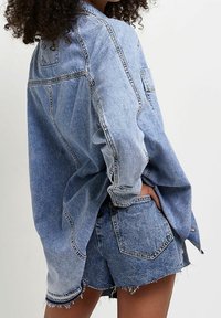 Person klädd i en överdimensionerad ljusblå denimjacka och matchande högmidjade denimshorts med fransar, med händerna i fickorna, mot vit bakgrund.