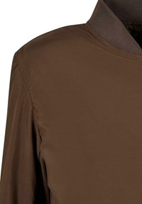 Tela marrón, ligera y con una textura suave, que presenta un cuello estructurado y detalles en las costuras, diseñada para una silueta ajustada.