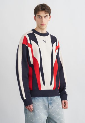 Młody mężczyzna w wielokolorowym swetrze Puma w odcieniach czerwieni, granatu i kremu oraz jasnoniebieskich dżinsach z subtelnym wzorem logo Puma na jednolitym tle.