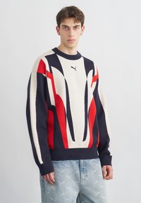 Giovane uomo che indossa un maglione Puma multicolore rosso, navy e crema e jeans azzurri con un sottile motivo a logo Puma su uno sfondo semplice.