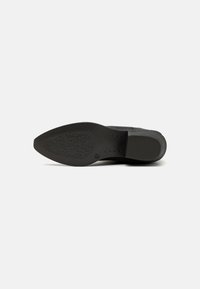 Semelle de chaussure noire en forme pointue, avec un motif de grip texturé et des bords latéraux lisses. Présente un petit logo imprimé près du talon.