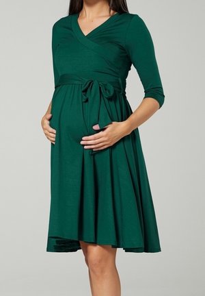 Femme enceinte portant une robe verte longueur genou avec des manches trois-quarts, tenant son ventre avec les deux mains.