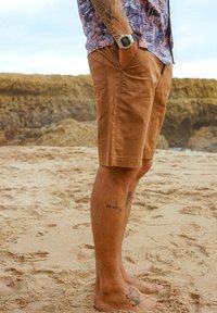 Shorts en coton marron avec poches latérales, offrant une coupe décontractée. Une personne se tient sur une plage de sable, montrant des tatouages sur ses jambes. Des empreintes de pas sont visibles dans le sable.