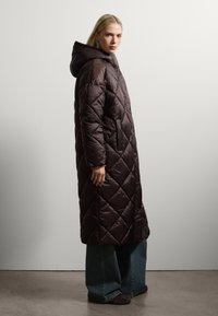 Cappotto imbottito marrone, trapuntato e con cappuccio. Presenta un motivo a forma di diamante, tasche laterali e una lunghezza lunga, abbinato a jeans a gamba larga.