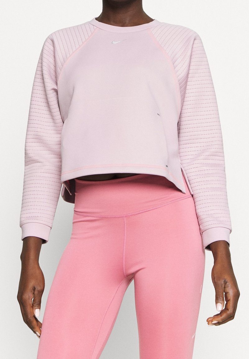 Femme portant un sweat-shirt matelassé à manches longues rose clair et un legging taille haute rose uni sur fond blanc.