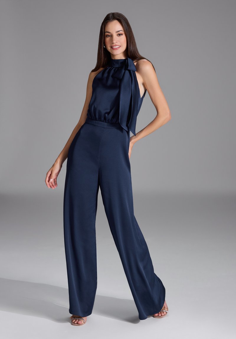 Combinaison bleu marine en satin avec col montant et détail de nœud, design à jambes larges et taille sans couture, mise en valeur par des talons à lanières.