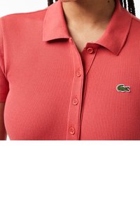 Polo en côte corail avec col, manches courtes et fermeture à boutons. Présente un petit logo crocodile brodé en vert.