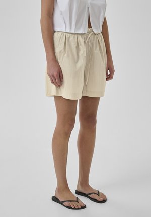 Frau trägt beige Shorts mit Kordelzug, weißes bauchfreies Oberteil und schwarze Flip-Flops, steht vor einem schlichten hellgrauen Hintergrund.