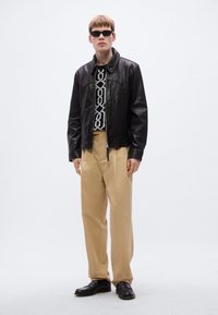 Μαύρο δερμάτινο μπουφάν πάνω από ασπρόμαυρο πουλόβερ με μοτίβο, συνδυασμένο με μπεζ παντελόνι και μαύρα παπούτσια, αξεσουάρ με γυαλιά ηλίου.