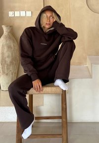 Kvinde i mørkebrun hættetrøje og bukser sidder på en træskammel, barfodet med hvide sokker, læner sig på hånden, beige struktureret væg og stor vase i baggrunden.