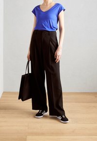 Kvinna i blå kortärmad topp, högmidjade svarta vida byxor, svarta sneakers och håller en svart tote-väska, stående på ett trägolv.