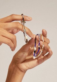 Zwei Perlenarmbänder werden in Händen gehalten. Das eine ist bunt mit blauen, schwarzen und weißen Perlen; das andere hat graue und braune Akzente.