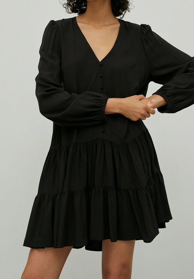 Robe noire avec un col en V, des manches longues bouffantes et des détails boutonnés. Elle possède une jupe à volants à étages avec un bord froncé et une texture de tissu douce.