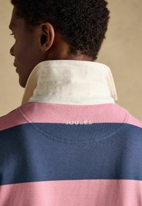 Personne aux cheveux courts et bouclés portant une chemise rayée rose et bleu marine avec un col blanc relevé, et "Joules" brodé dans le dos.