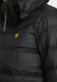 Lähikuvassa musta puhvijakku, jossa on huppu, etuvetoketju ja pieni keltainen "RAW"-logo rinnassa.