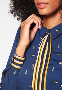 Mörkblå skjorta med fågelmönster, med en knäppt krage och randiga manschetter i gult och marinblått. Slen textur, skräddarsydd passform.