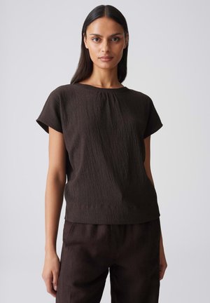 Donna con capelli lunghi e scuri che indossa una maglia a maniche corte marrone scuro testurizzata e pantaloni marrone scuro abbinati, in piedi davanti a uno sfondo chiaro uniforme.