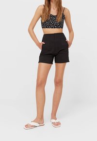 Pantaloni shorts neri a vita alta con tasche, caratterizzati da una texture liscia e un design sartoriale, abbinati a un top corto nero con motivo floreale e sandali bianchi.