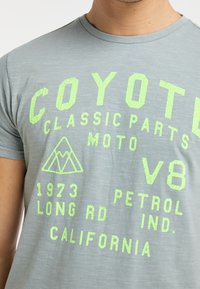 Šedé tričko s krátkým rukávem, zdobené prasklým neonově zeleným textem a grafikou, včetně nápisu "COYOTE" a designu trojúhelníku. Měkký, lehký materiál.
