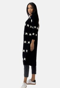 Gilet noir en tricot avec des motifs d'étoiles blanches, à manches longues et à coupe décontractée, porté sur un jean noir coupé et des baskets blanches.