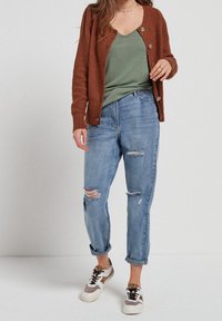 Cardigan en maille couleur rouille, haut vert en v, jean boyfriend clair déchiré, et baskets multicolores avec des motifs.