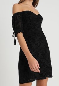 Vestido negro con hombros al descubierto, mangas abullonadas, textura floral, corsé ajustado y falda acampanada. Detalle de cordón en las mangas.