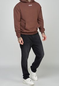 Felpa marrone con tasca frontale e logo bianco, abbinata a jeans scuri e sneakers bianche. Materiale misto cotone, vestibilità rilassata.