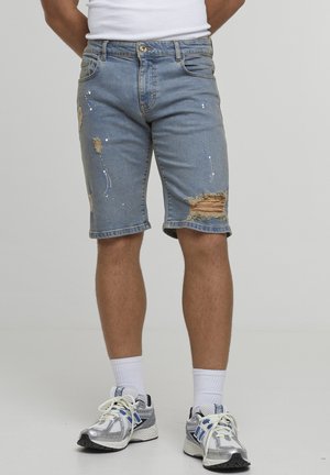 Hombre con pantalones cortos de mezclilla desgastados con salpicaduras de pintura, calcetines blancos y zapatillas de correr plateadas, de pie contra un fondo liso.