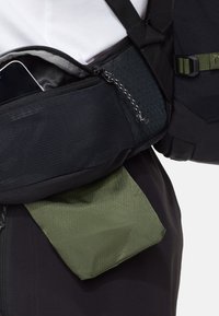 Nahaufnahme einer schwarzen Bauchtasche mit einer teilweise geöffneten Reißverschlusstasche, die ein Smartphone hält, getragen über dunkler Hose und weißem Oberteil.