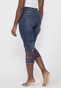 Femme portant un jean court bleu foncé avec une broderie florale près de l'ourlet et des sandales à talons hauts transparentes, debout devant un fond uni.