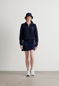 Sötétkék, cipzáras pulóver ribbed ujjakra, hozzáillő, oldalsávos szoknyával párosítva. Sötétkék virett kalap és fehér sneaker cipő mellett viselve.