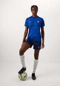 Blue soccer jersey with pattern and crest, paired with navy shorts. Black soccer shoes and white socks, standing on a patterned soccer ball.