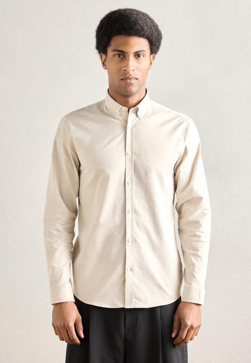 Μπεζ πουκάμισο με κουμπιά, κατασκευασμένο από υφή υφάσματος, με γιακά, μακριά μανίκια και στενή εφαρμογή με ευθεία ραφή στο τελείωμα.