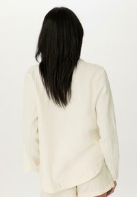 Světle krémová, texturovaná košile s volným střihem a dlouhými rukávy. Zaoblený lem a jemný vzor. Zadní pohled, s částečně viditelnými vlasy.