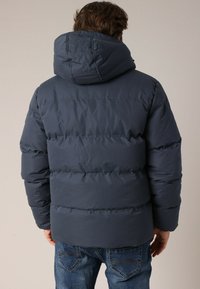 Veste matelassée bleu marine avec capuche, dotée d'un design quilté, de poignets élastiques et d'une coupe décontractée. Portée avec un jean bleu.