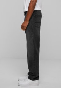 Black denim jeans with a straight-leg cut, five pockets, and minimal stitching, paired with white sneakers. Side view shows fit and design.