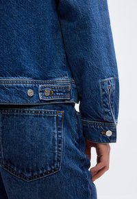 Κοντινό πλάνο ατόμου που φοράει μπλε τζιν μπουφάν και τζιν παντελόνι, δείχνοντας την πίσω τσέπη, τη μανσέτα του μπουφάν και το χέρι που ακουμπά κοντά στο ισχίο.