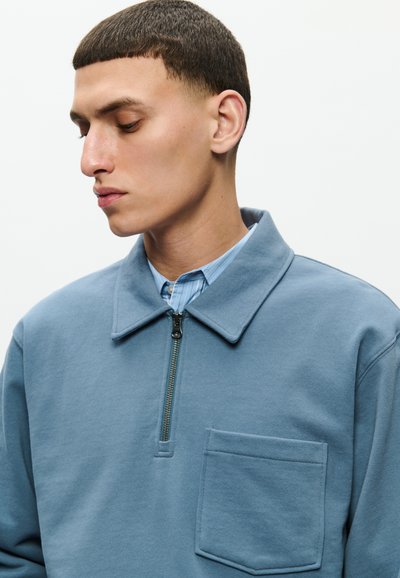 Young man with short dark hair looking down, wearing a blue zip-up shirt layered over a striped collared shirt, isolated on white background.