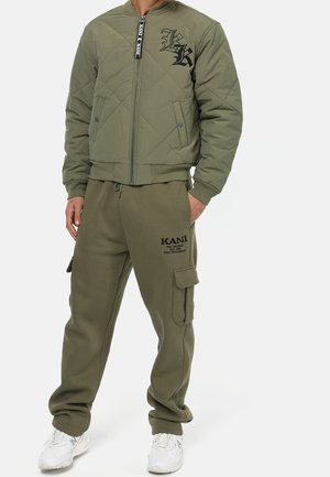 Mann trägt olivgrüne gesteppte Bomberjacke und passende Cargo-Sweathosen mit schwarzen Logos, kombiniert mit weißen Turnschuhen.