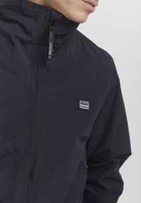 Giacca leggera nera con colletto alto e chiusura a zip. Presenta una piccola toppa con logo sul lato sinistro e un tessuto liscio e strutturato.