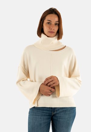 Giovane donna con capelli castani fino alle spalle indossa un maglione a collo alto beige a coste con maniche larghe e jeans blu, in posizione neutra.